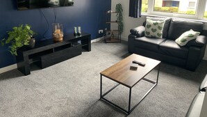 Una sala de estar con un televisor montado en la pared, un sofá negro con almohadas estampadas con corazones, una mesa de centro de madera y una alfombra gris.