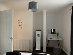 Ein minimalistisches Schlafzimmer mit einer weißen Tür, einem Heizkörper, einem großen Spiegel, einem weißen Nachttisch mit Pflanze und einem motivierenden Zitat an der Wand.