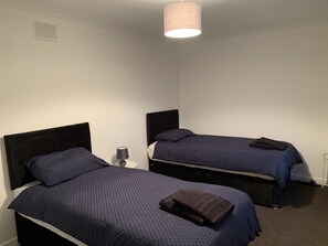 A room with two single beds, each with a headboard, a bedside table with a lamp, and a folded towel on the bed.