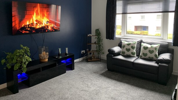 Una sala de estar con un televisor que muestra una chimenea, una pared azul oscuro, un sofá gris con cojines estampados de corazones y una ventana con persianas.