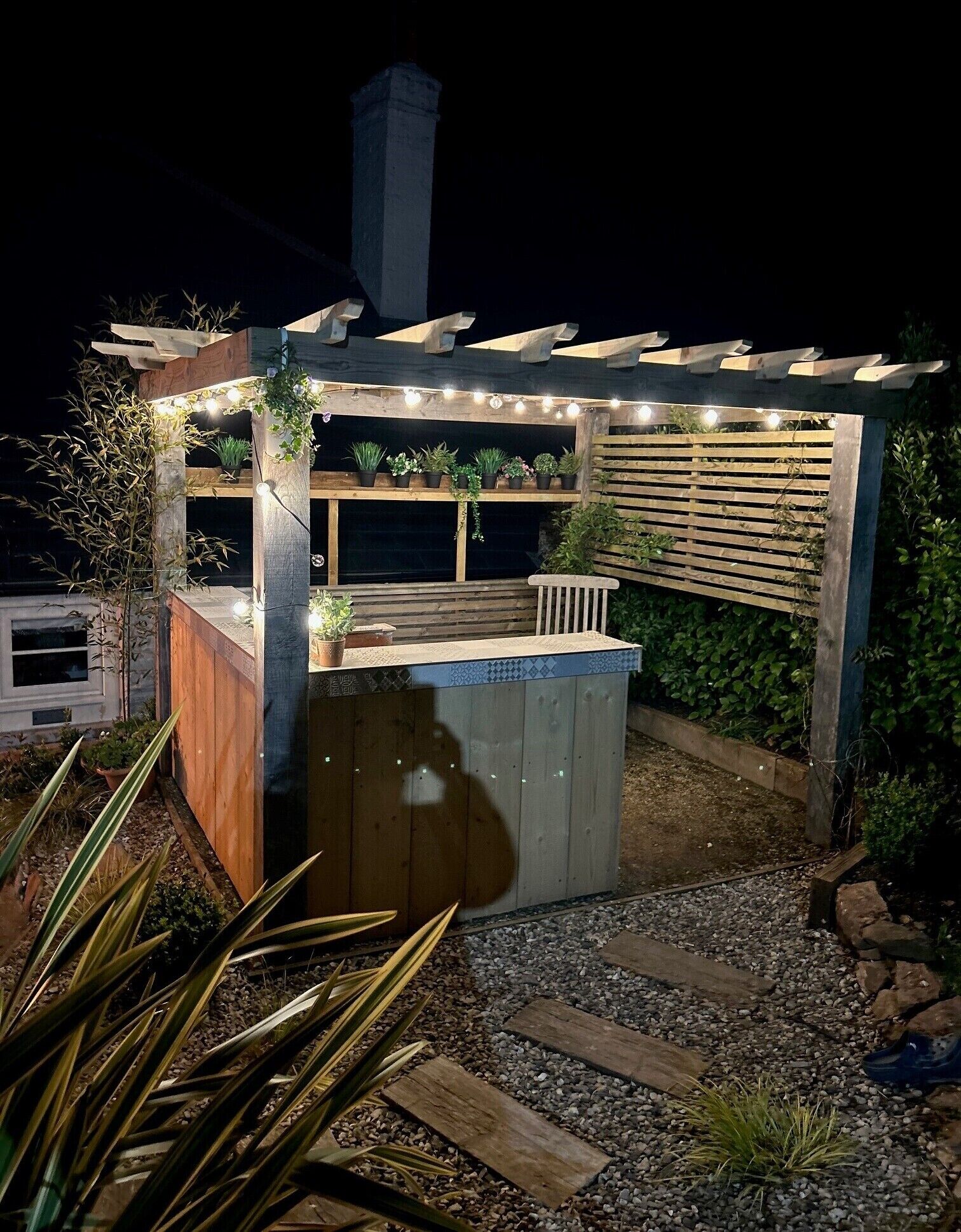 A wooden pergola with string lights, a wooden bench, and potted plants.