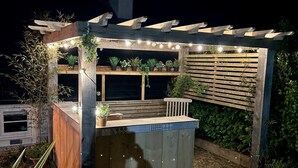 A wooden pergola with string lights, a wooden bench, and potted plants.