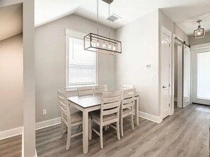 Un coin repas avec une table et des chaises blanches, un luminaire suspendu et une fenêtre avec des stores.