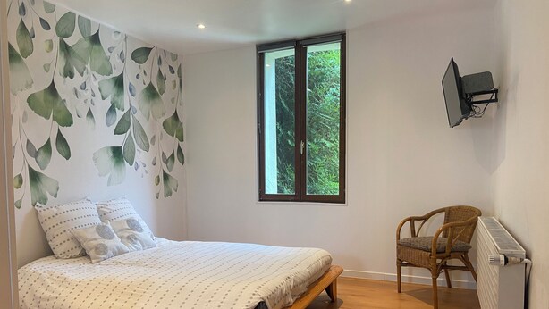 A bedroom with a bed, a chair, a radiator, and a window with a view of greenery.