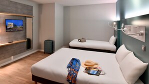 A hotel room with a large bed, a smaller bed, a TV mounted on the wall, a desk with a chair, and a trash bin. - Ibis Budget Saint Nazaire Centre Gare (Saint-Nazaire)