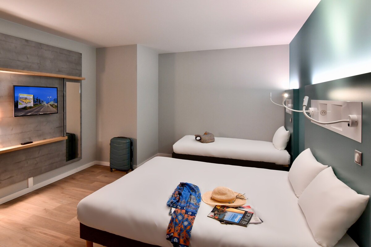 Une chambre d’hôtel avec un grand lit, un lit plus petit, un téléviseur fixé au mur, un bureau avec une chaise et une poubelle.