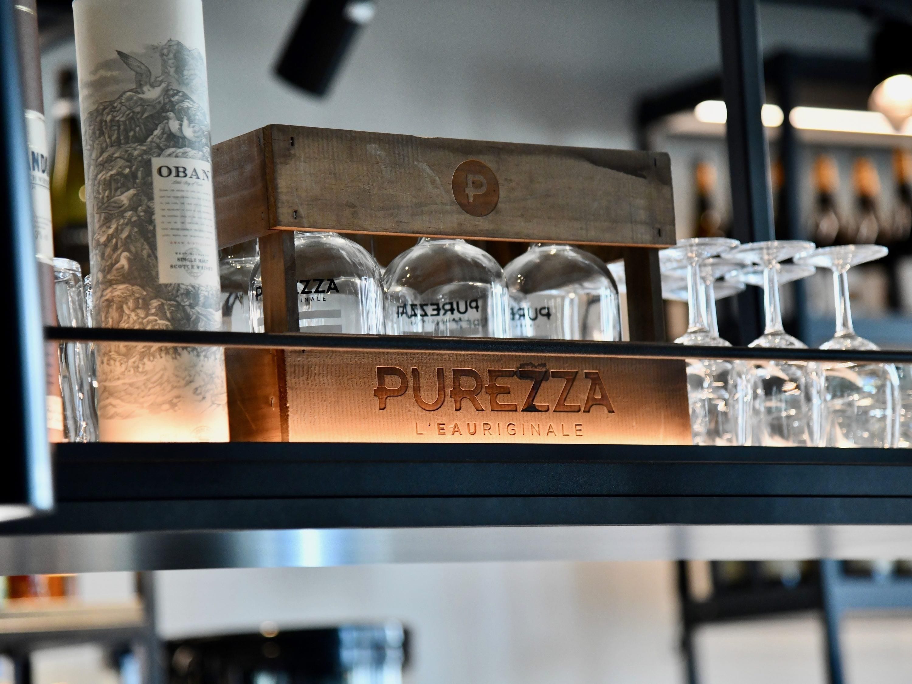 A bar area with a wooden display case labeled 'PUREZZA L'EAU ORIGINALE' containing glasses and bottles.