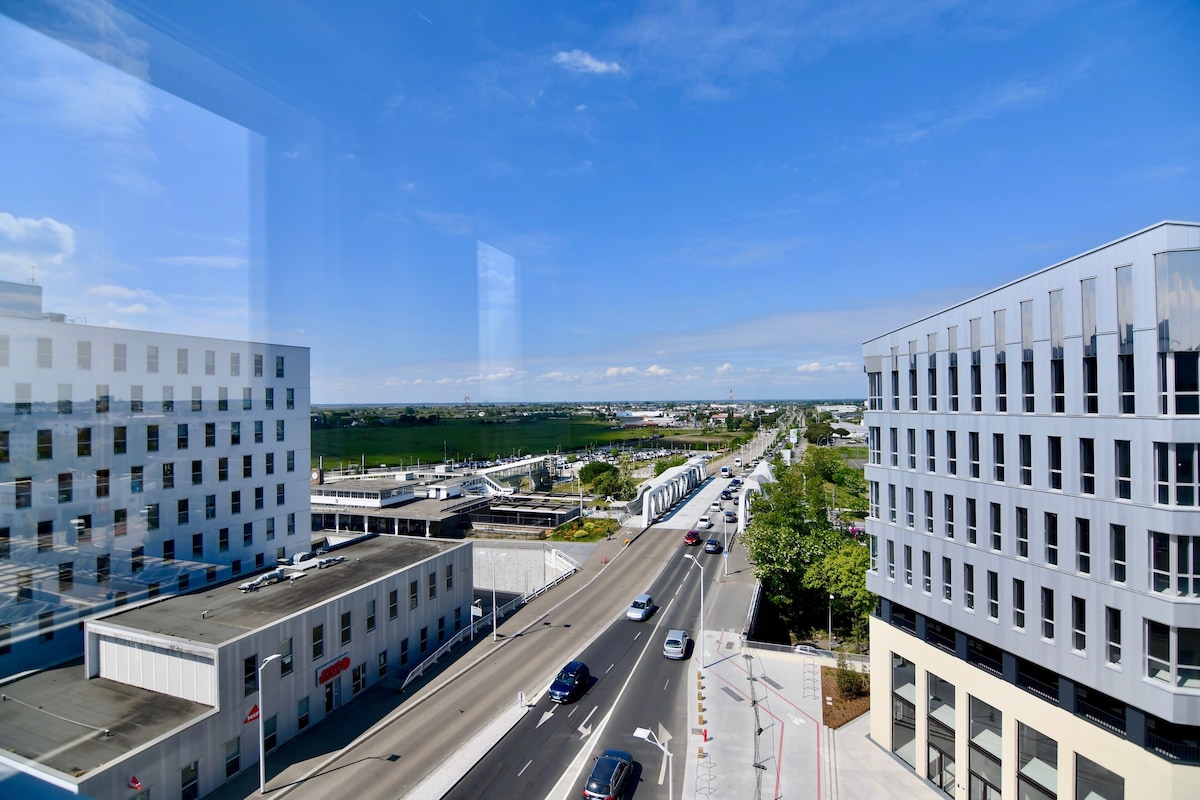 Un bâtiment moderne doté d’un toit plat, de grandes fenêtres et offrant une vue sur une route où circulent des voitures, avec une étendue d’eau en arrière-plan.