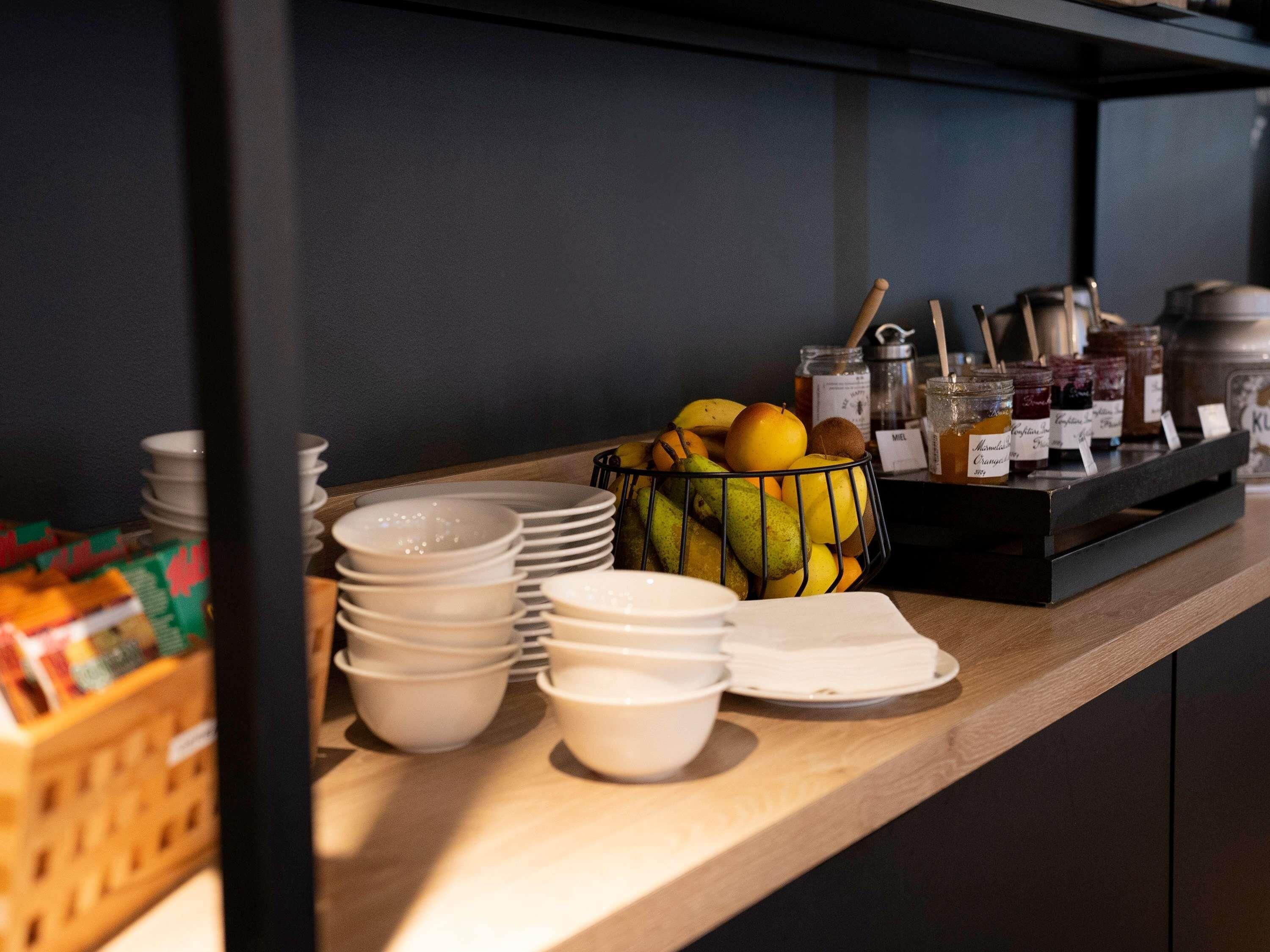 A buffet setup with stacked white plates, bowls, and a fruit basket containing apples, pears, and bananas.