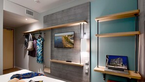 A modern hotel room with a bed, a TV mounted on a grey wall, a laptop on a wooden shelf, and a wardrobe with hanging clothes. - Ibis Budget Saint Nazaire Centre Gare (Saint-Nazaire)