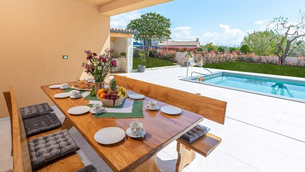 A dining area with a poolside view, featuring a wooden table set for breakfast, a bench, and a view of a swimming pool and garden.