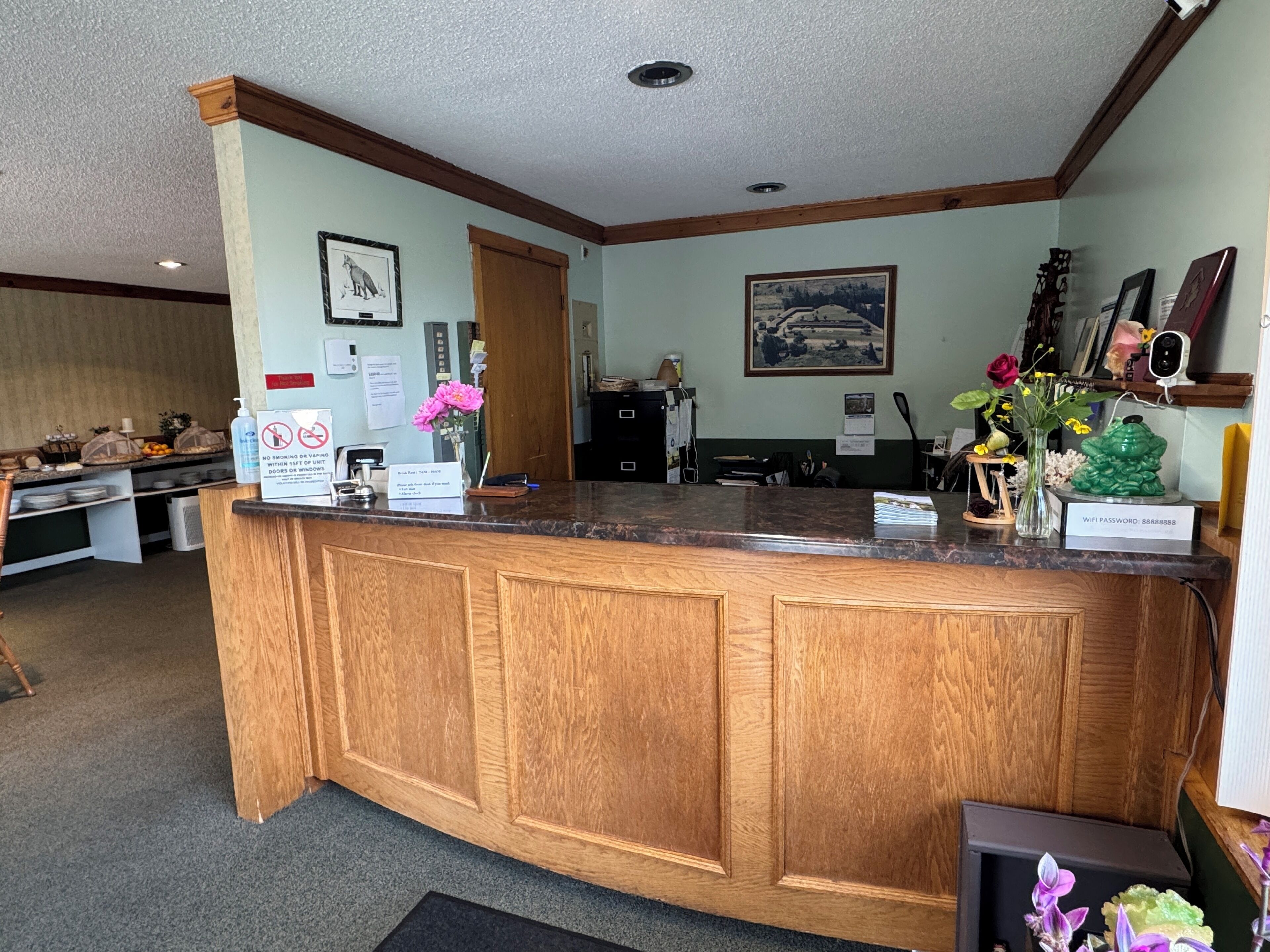 A reception desk with a wooden counter, a no-smoking sign, a framed picture, and a vase with flowers.