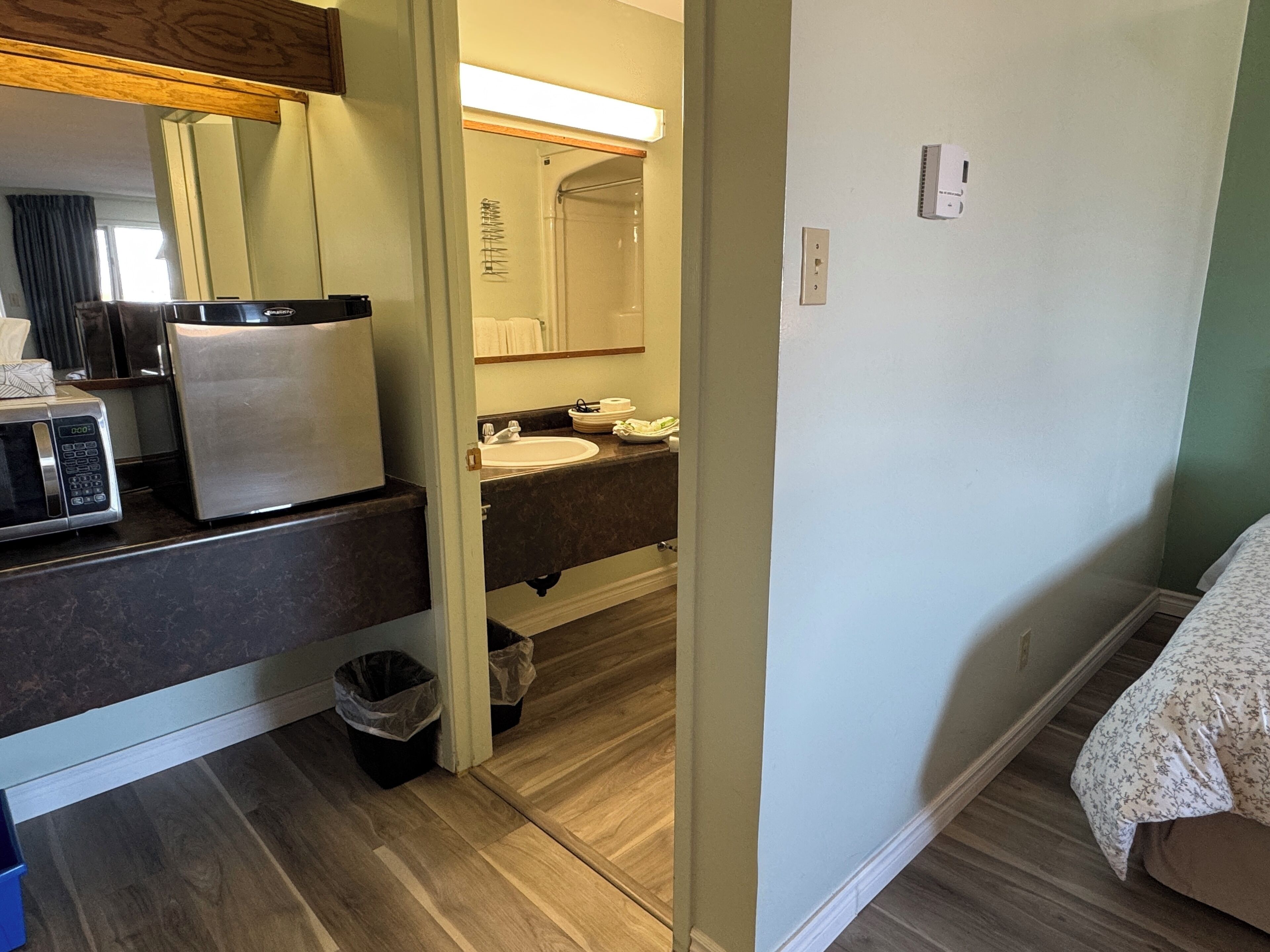 A compact hotel room with a kitchenette, a bathroom with a mirror and sink, and a bed with a floral bedspread.