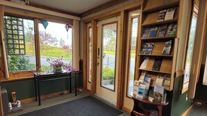 Un área de recepción con un mostrador de madera, un felpudo negro, una estantería de madera con folletos y vistas al exterior a través de una puerta de vidrio.