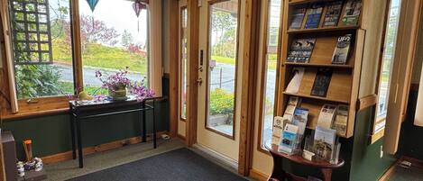 A reception area with a wooden reception desk, a black mat, a wooden display shelf with brochures, and a view of the outdoors through a glass door.