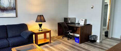A living room with a sofa, a coffee table, a lamp, a desk with a computer and a microwave, and a painting on the wall.