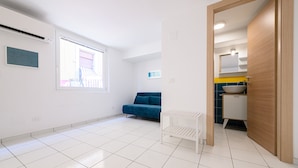 A spacious room with white tiled flooring, a blue sofa, a white radiator, and a wooden door leading to a bathroom with a sink and mirror. - Via S Liborio - 6 Piano (Naples)