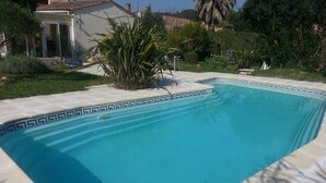 Une piscine rectangulaire bordée de carreaux, entourée d’un jardin où poussent différentes espèces de plantes, avec une maison en arrière-plan.