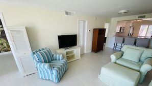A living room with a striped sofa, a light-colored sofa, a TV on a white stand, and a kitchen area in the background.