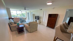 A spacious living room with a sofa, armchairs, a coffee table, and a TV. There is a wooden cabinet and a door leading to another room.