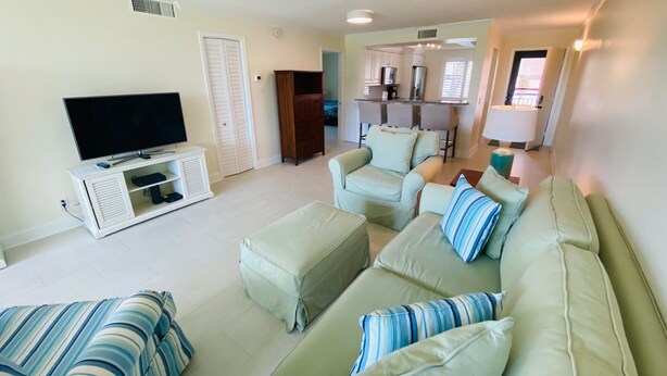 A living room with a TV, sofa, and ottoman. There is a kitchen area in the background.
