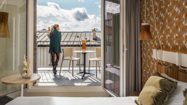 Habitación de hotel con balcón, cama, una mesita con un florero y pared con estampado.