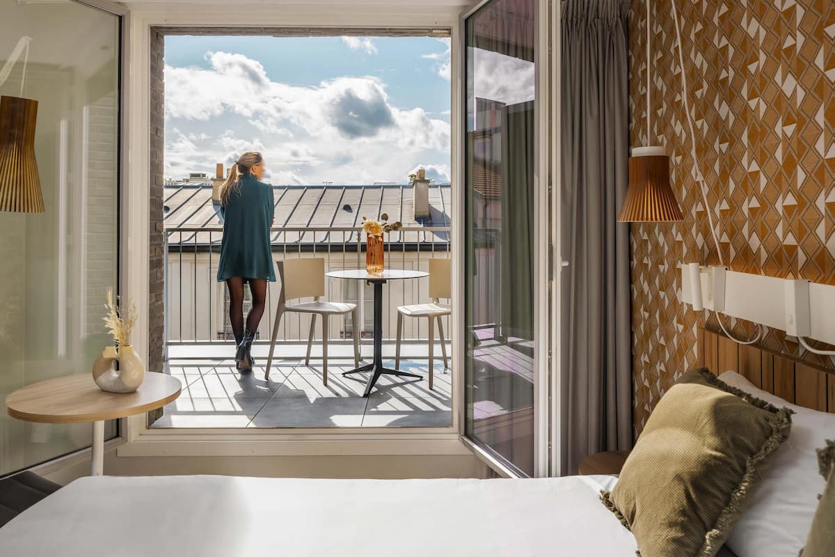 Une chambre d’hôtel avec un balcon, un lit, une petite table avec un vase et un mur décoré de motifs.