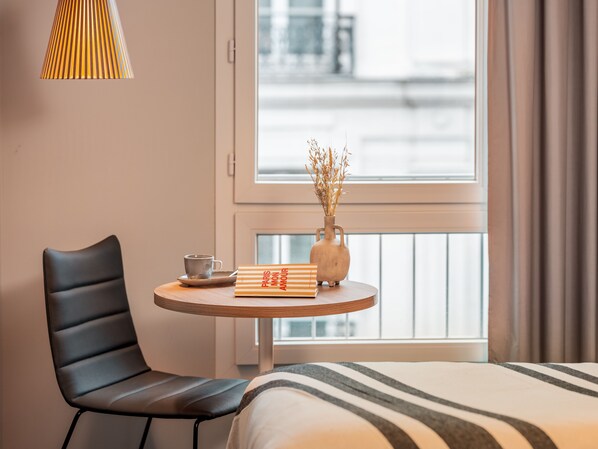 Une chambre d’hôtel avec un lit, une table ronde ornée d’un vase contenant des plantes séchées, une chaise et une lampe.