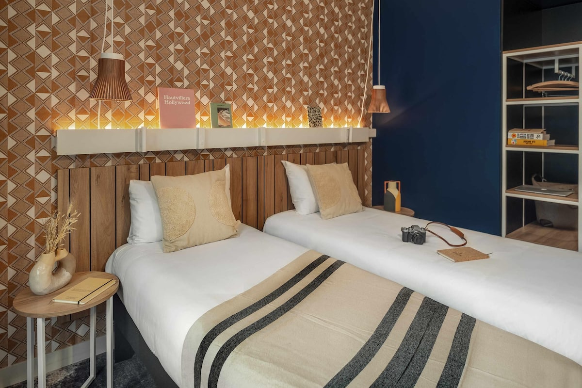 Une chambre d’hôtel avec un papier peint à motifs, une tête de lit en bois, deux lits avec une literie à rayures, une table de chevet avec un appareil photo, et une étagère avec des livres et une lampe.
