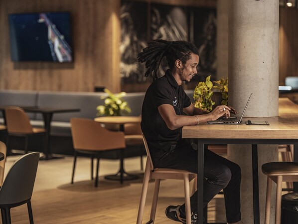 Eine Person sitzt an einem Tisch mit einem Laptop in einem Raum mit Holzboden, Stühlen und einem an der Wand befestigten Fernseher.