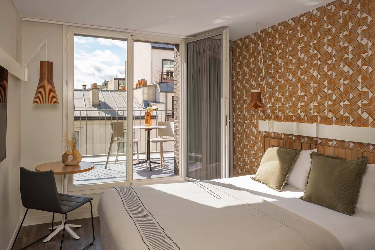 Habitación de hotel con cama, una mesita con sillas, un balcón con vistas a edificios y una pared con diseño.