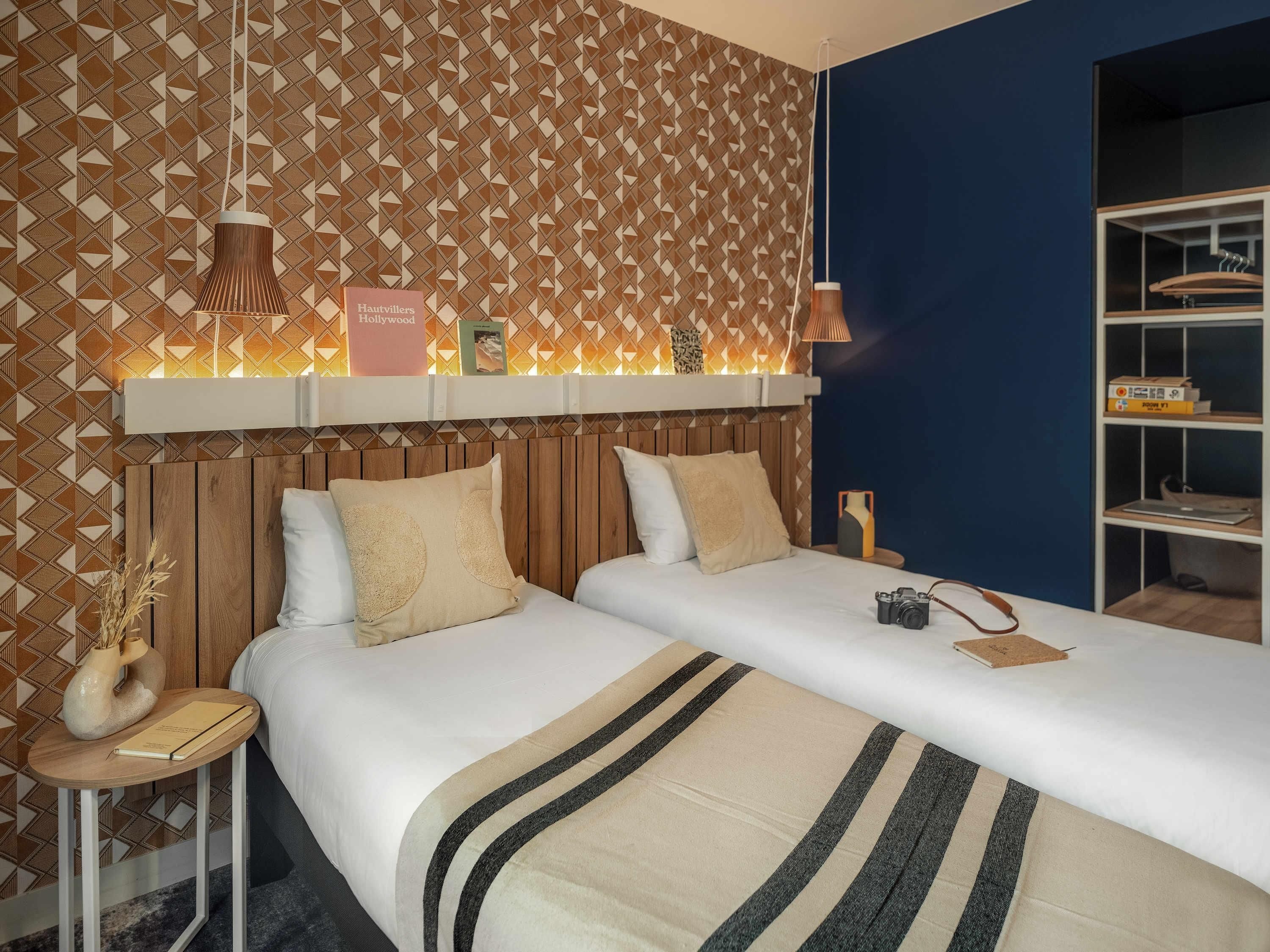 A hotel room with a patterned wallpaper, a wooden headboard, two beds with striped bedding, a bedside table with a camera, and a shelf with books and a lamp.
