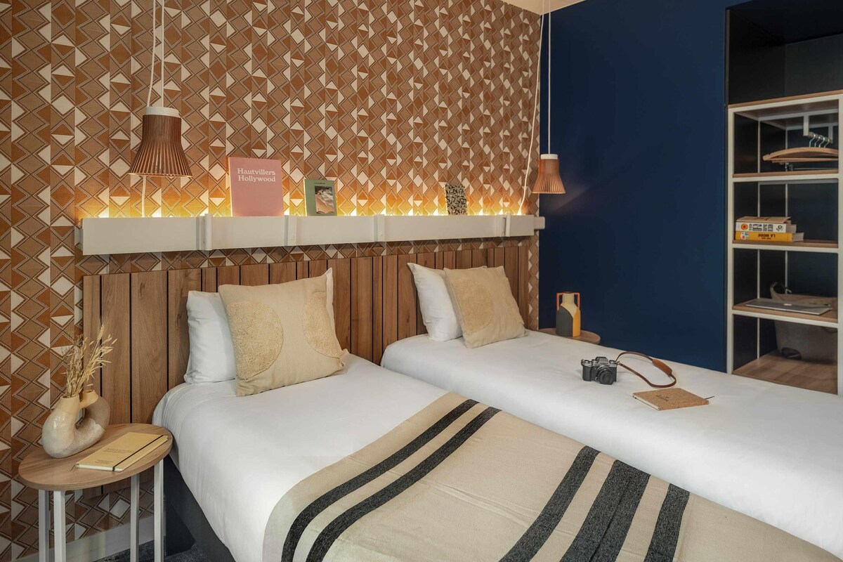 Habitación de hotel con papel tapiz estampado, cabecera de madera, dos camas con ropa de cama a rayas, mesita de noche con una cámara y estante con libros y una lámpara.