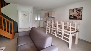Sala de estar com um sofá cinza, cadeiras de madeira, escada e uma área de cozinha com armários e geladeira.