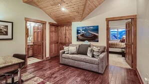Una sala de estar acogedora con techo de madera, un sofá gris con cojines, un cuadro en la pared y una ventana con vistas a un edificio.