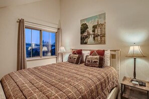A bedroom with a large bed, two bedside lamps, a painting of a cityscape, and a window with curtains.