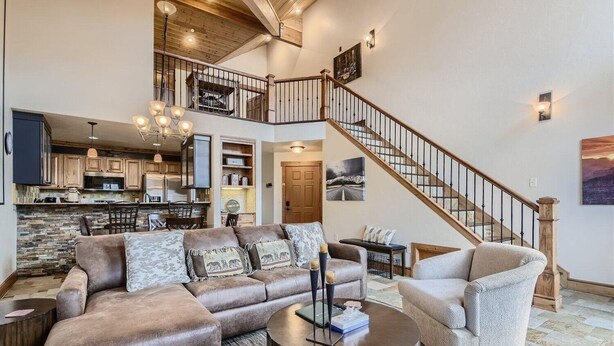 Un amplio salón con un sofá de cuero marrón grande, un sillón beige y una mesa de centro. Una escalera con barandillas de madera conduce a un nivel superior.
