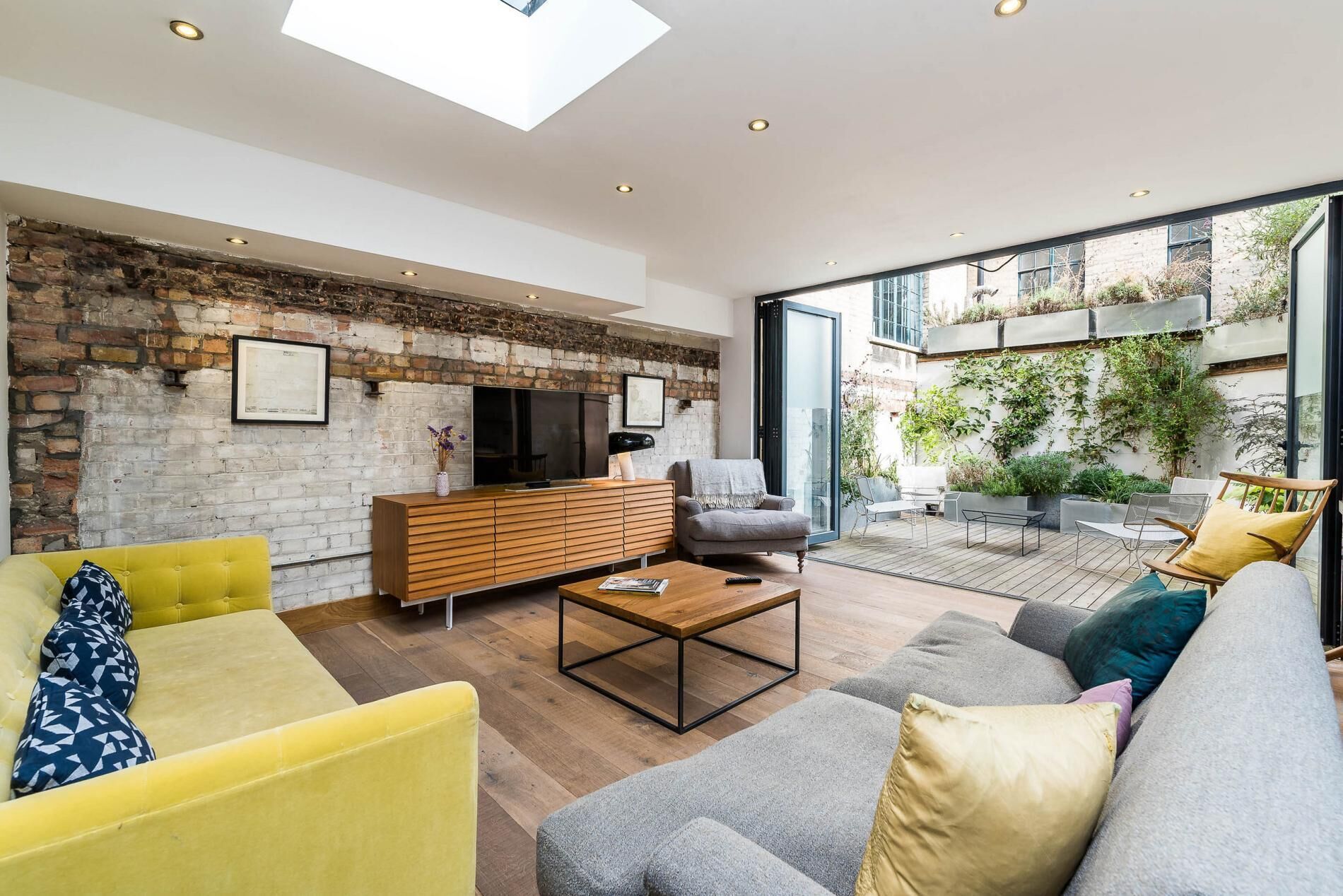 A spacious living area with a brick wall, a large TV, and a cozy seating arrangement including a yellow sofa and a grey couch.