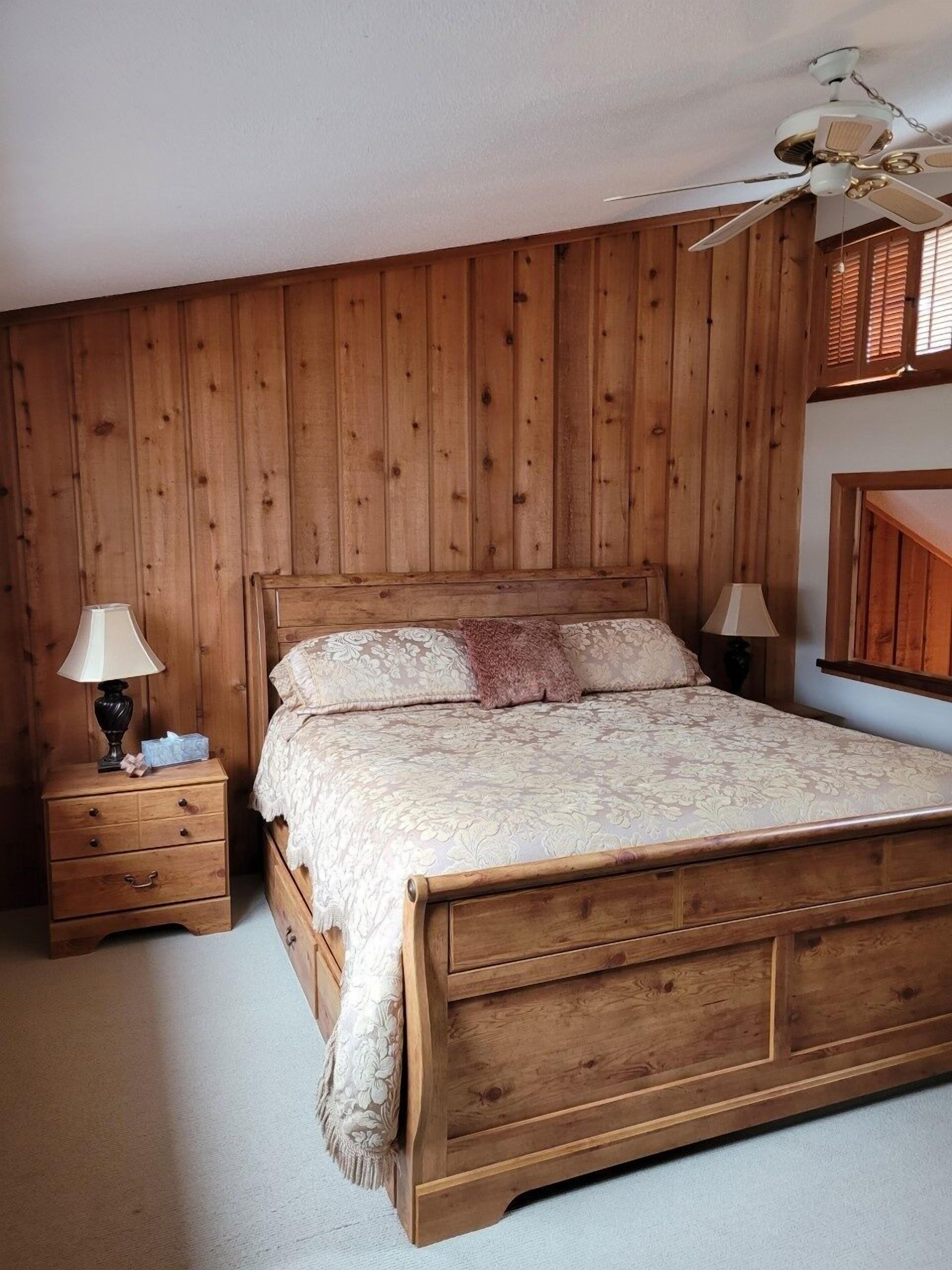 Une chambre à coucher aux murs lambrissés, avec un lit, deux tables de chevet et un ventilateur de plafond.