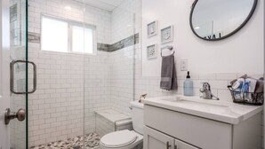 Une salle de bain avec une douche à parois de verre, des murs recouverts de carreaux blancs, des toilettes blanches, un meuble-lavabo avec un miroir et un porte-serviettes.