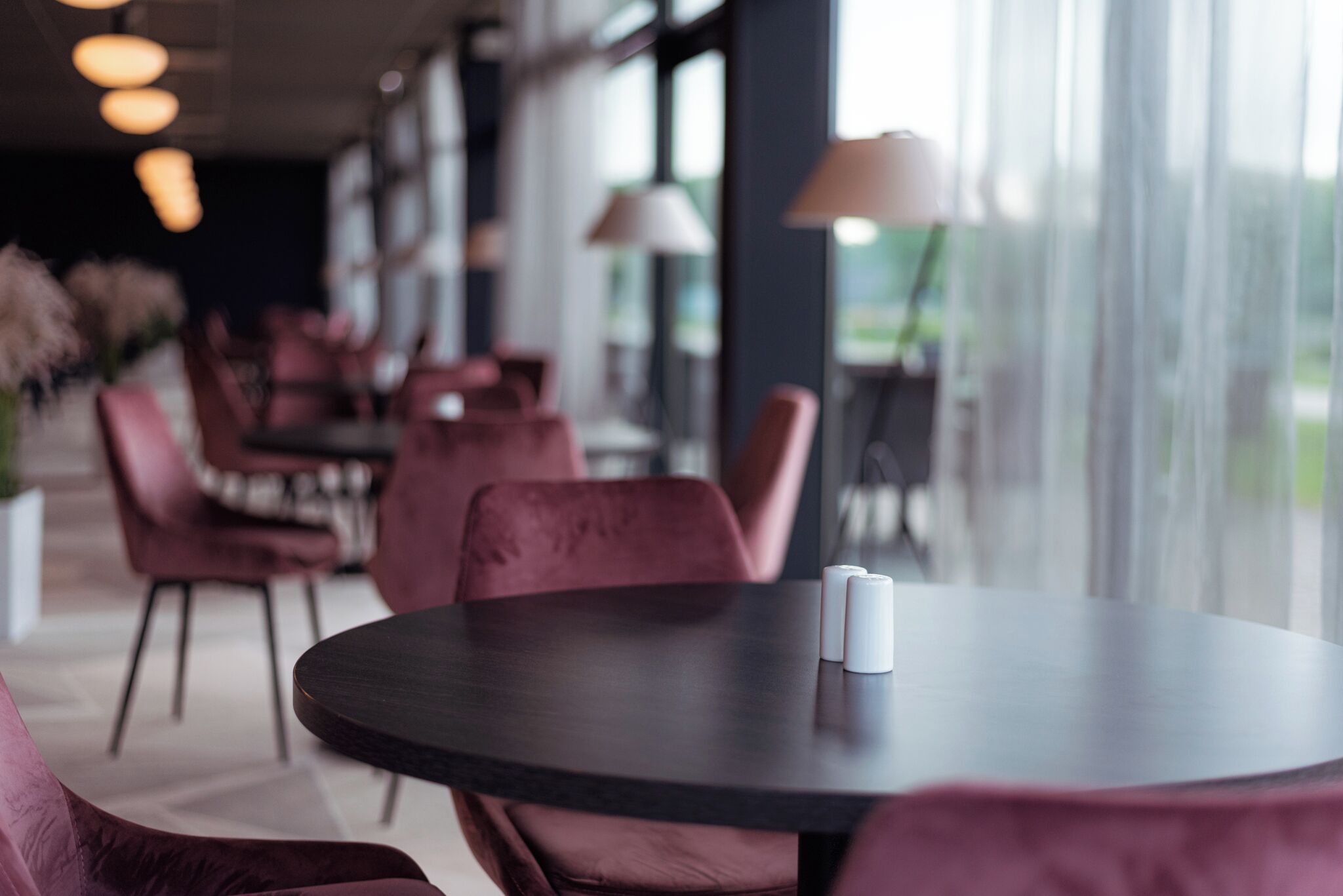 Interior moderno de restaurante con mesas redondas y sillas rojas cómodas, ventanas amplias y lámparas colgantes.
