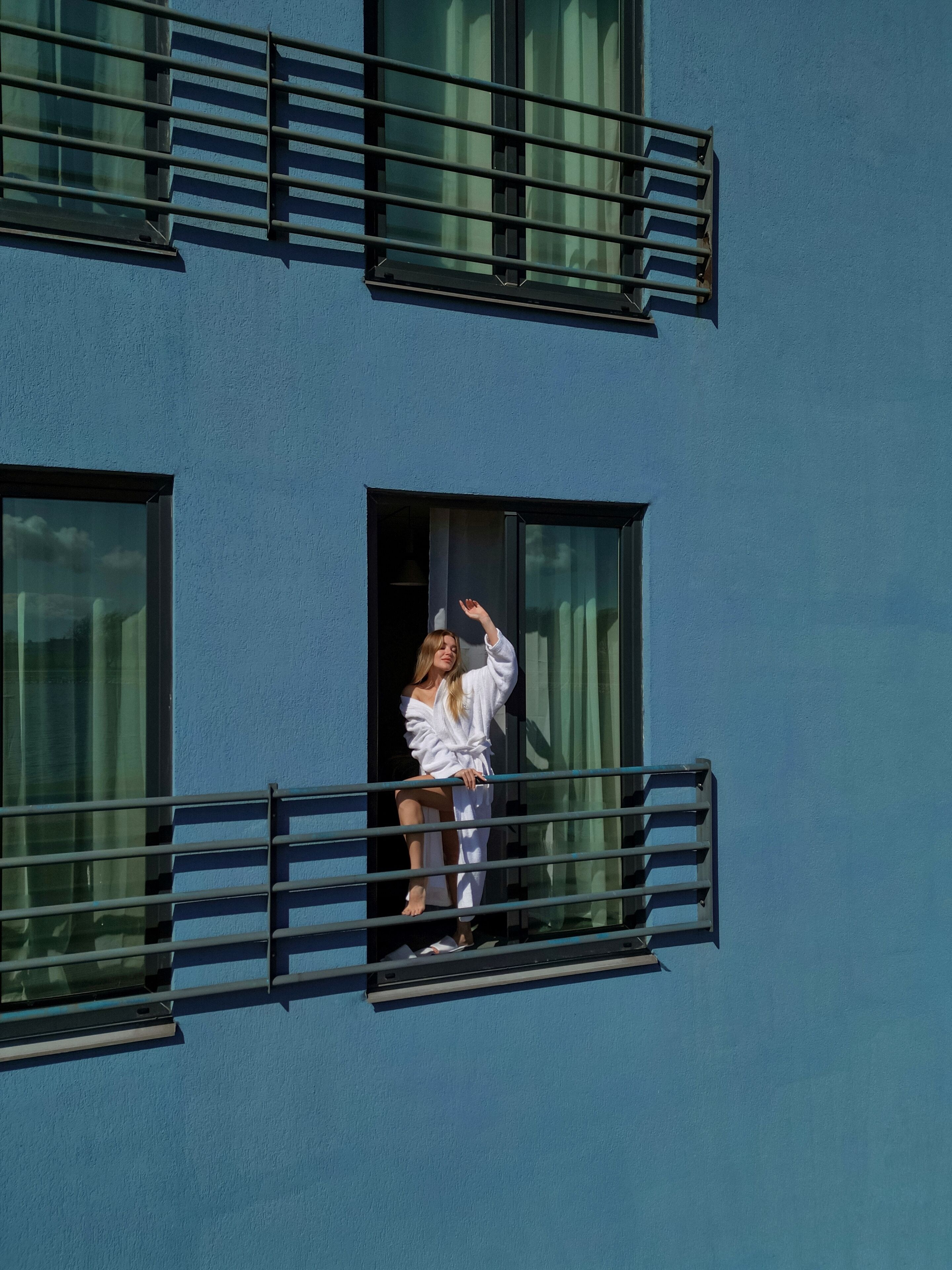 Una persona con un abito bianco in piedi su un balcone con ringhiere metalliche, che guarda un edificio con ampie vetrate e facciata blu.