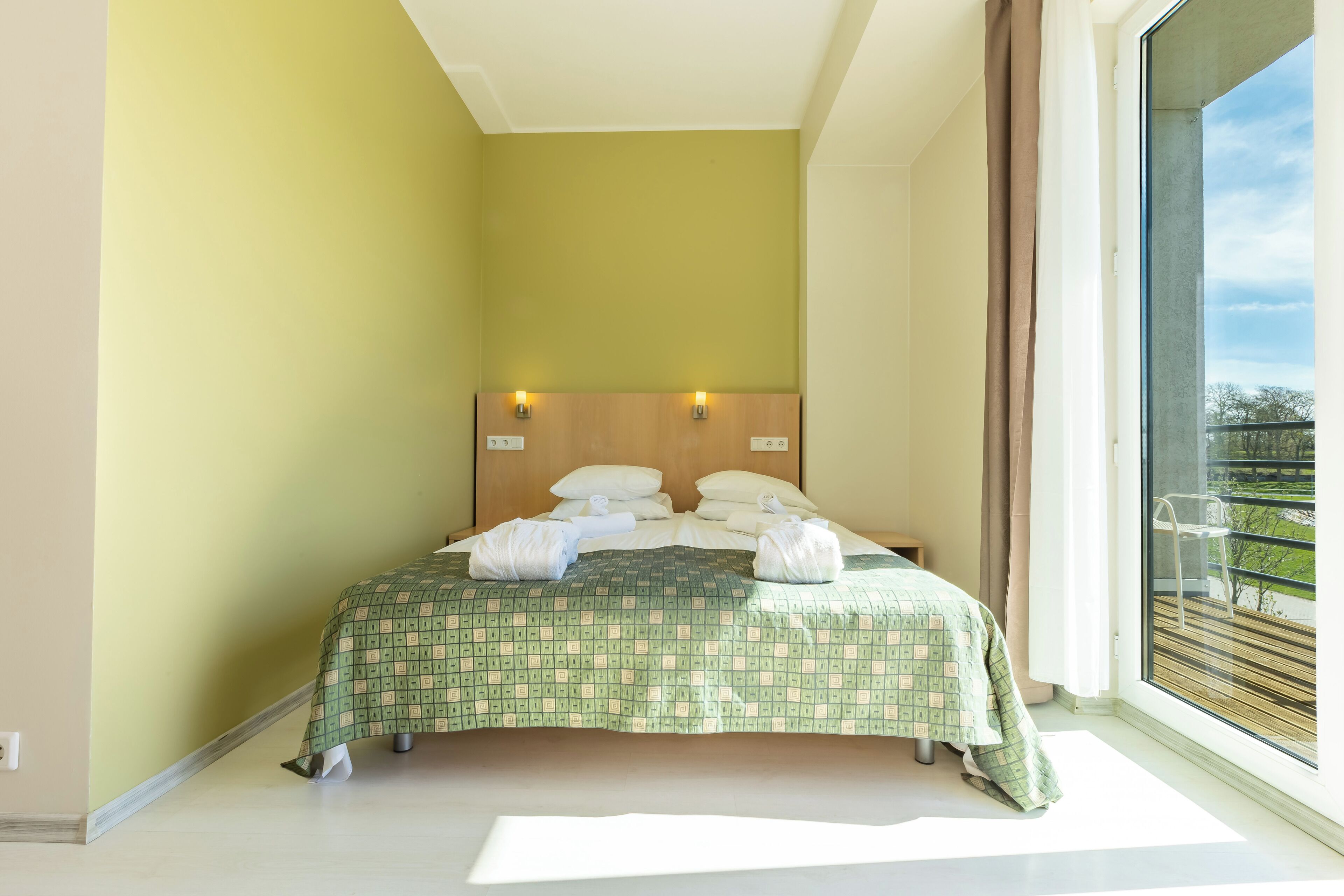 A hotel room with a large bed, a wooden headboard, two wall-mounted lights, and a view of the outdoors through a large window.