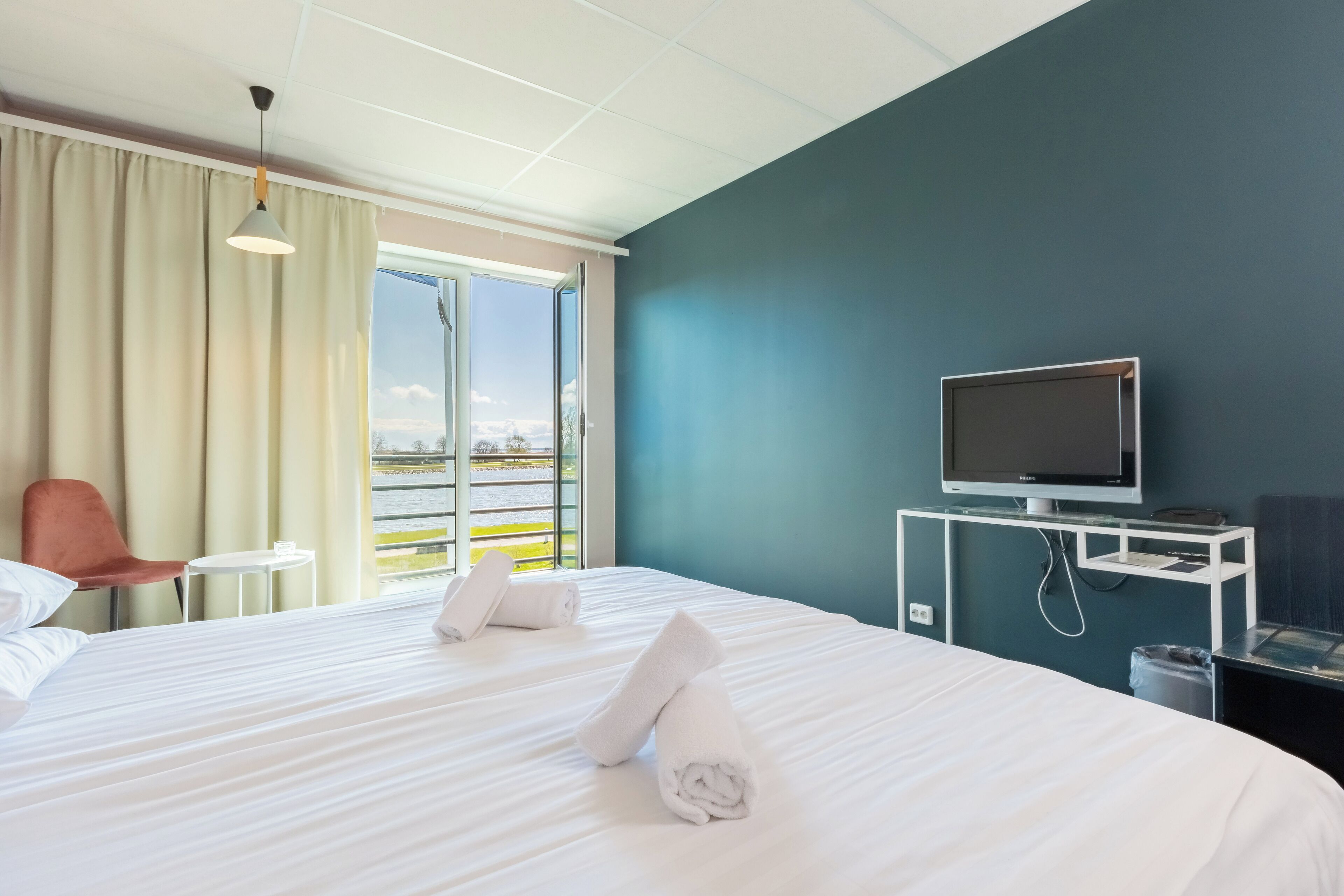 A hotel room with a bed, a TV on a stand, a chair, a small table, and a view of the outside.