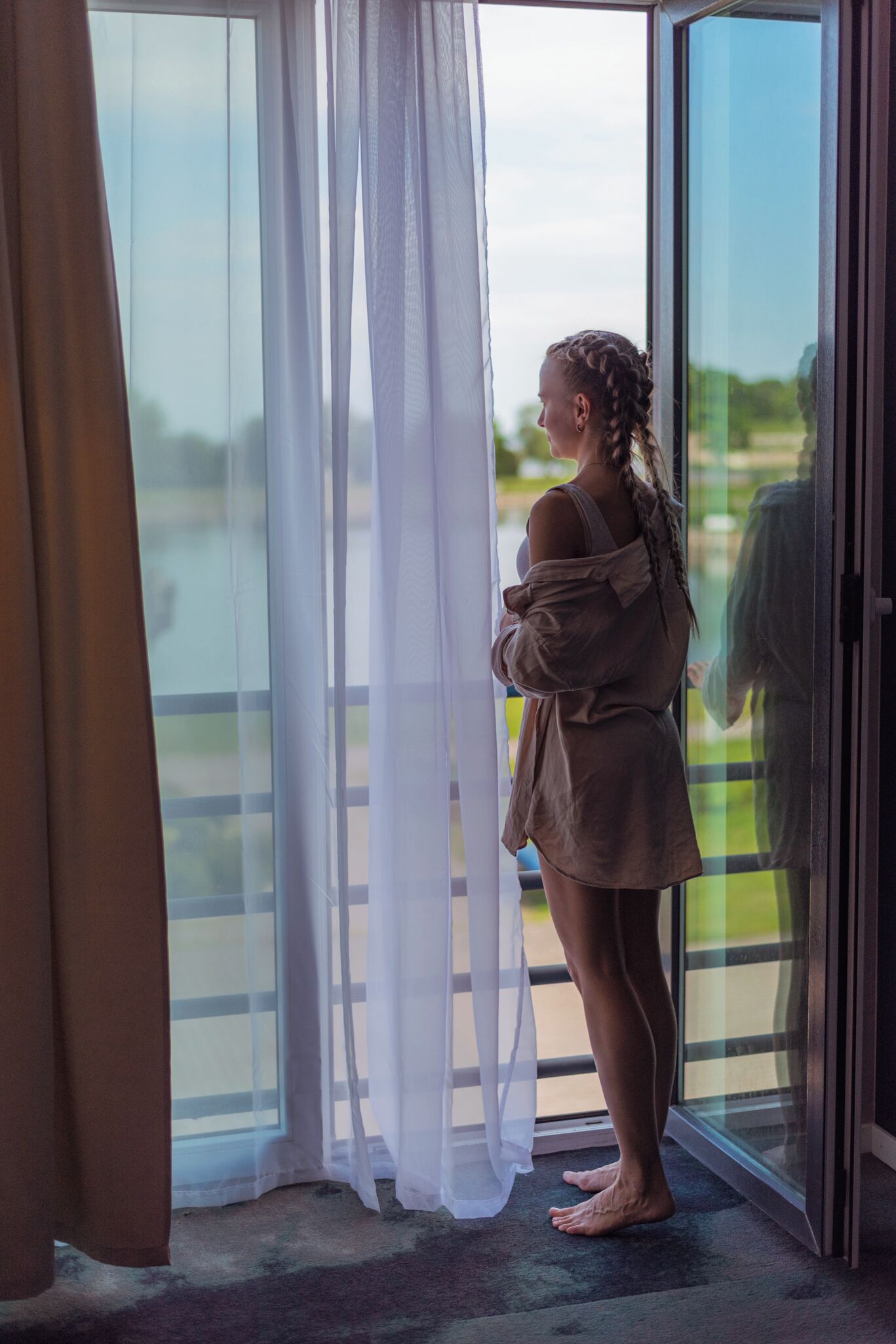 A person standing by a glass door with sheer curtains, looking outside.