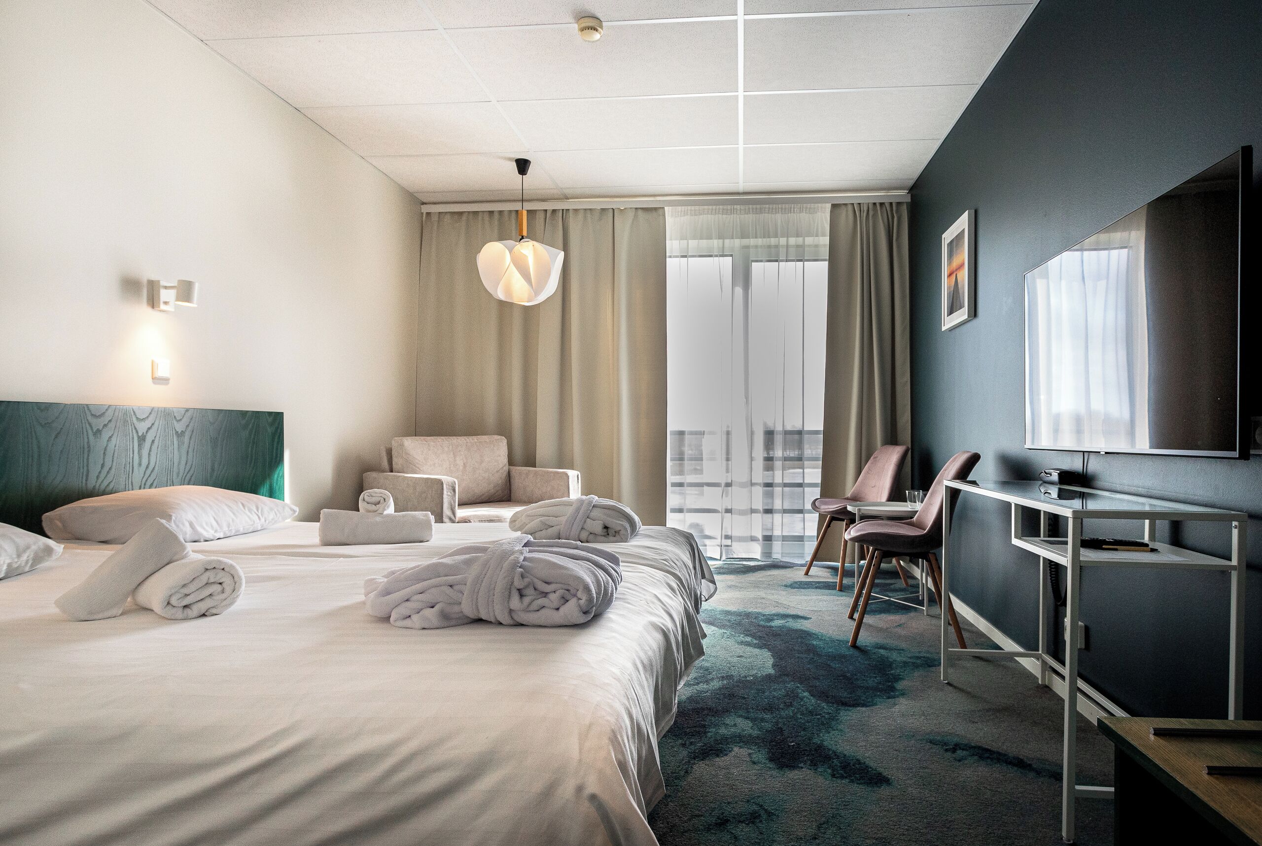A modern hotel room with a large bed, a desk, a chair, and a TV.