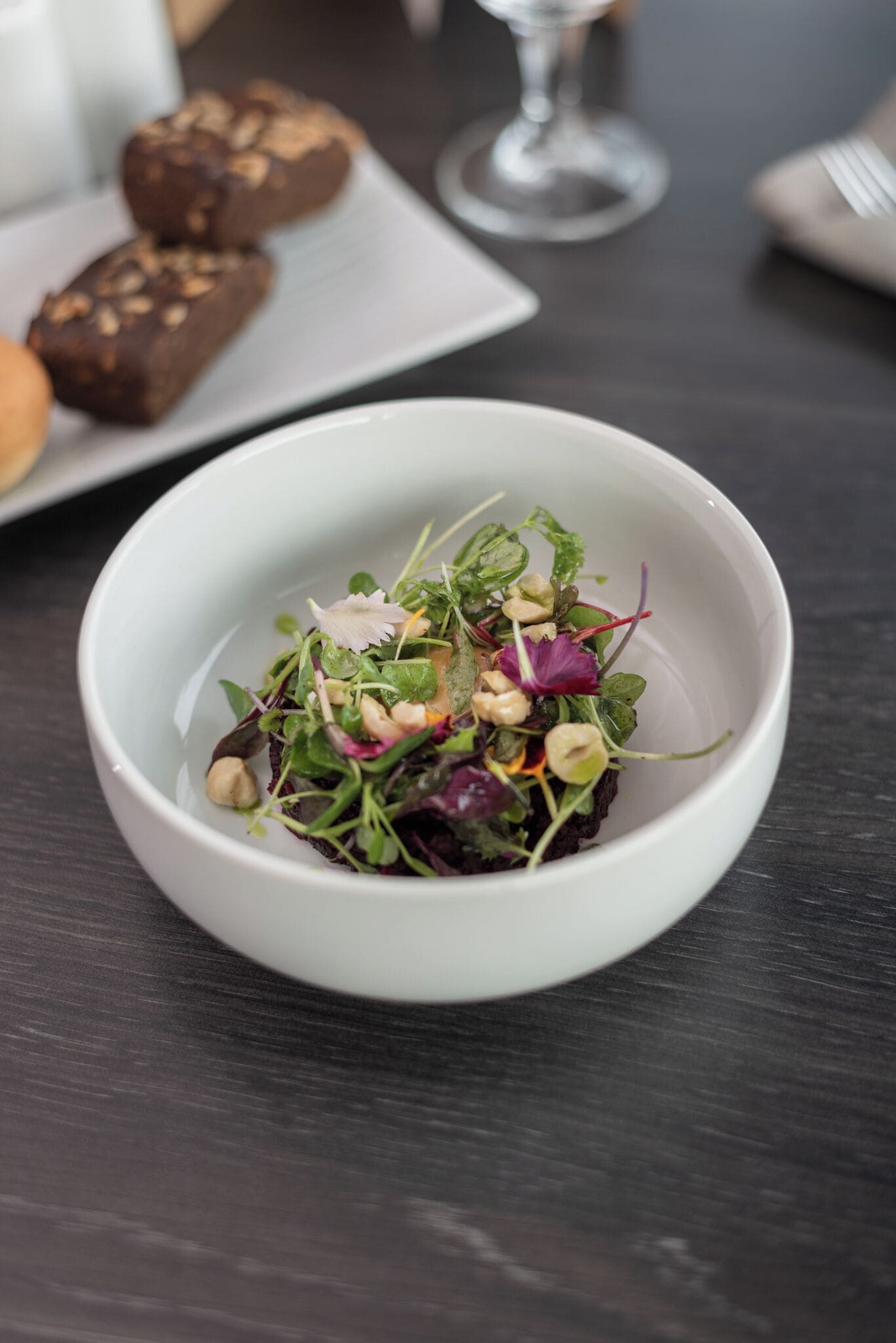 Eine weiße Schüssel mit Salat, verschiedenen Blattsalaten und Nüssen auf einem dunklen Holztisch.
