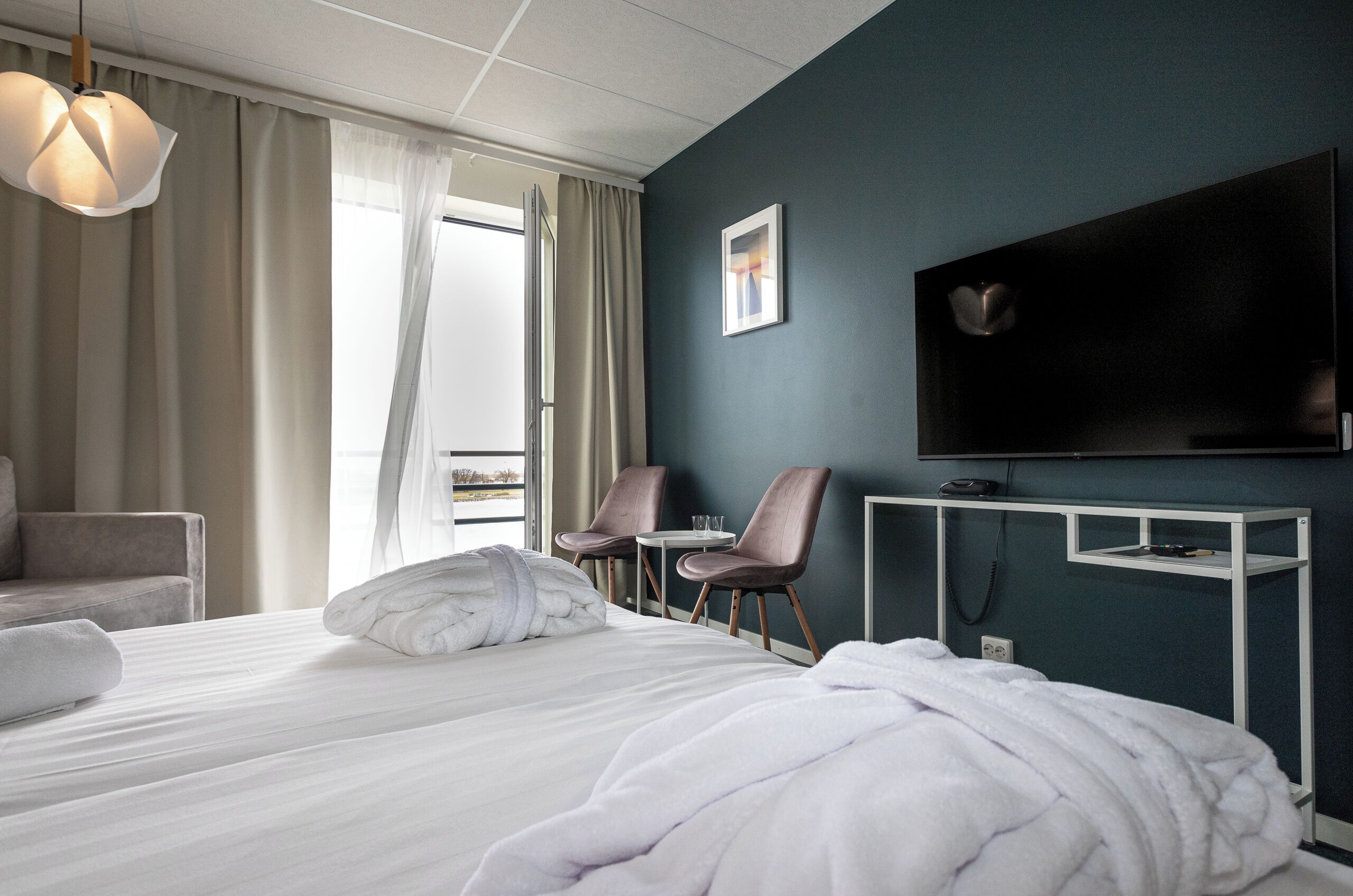 Une chambre d’hôtel avec un lit, deux chaises, une télévision et une fenêtre avec des rideaux.