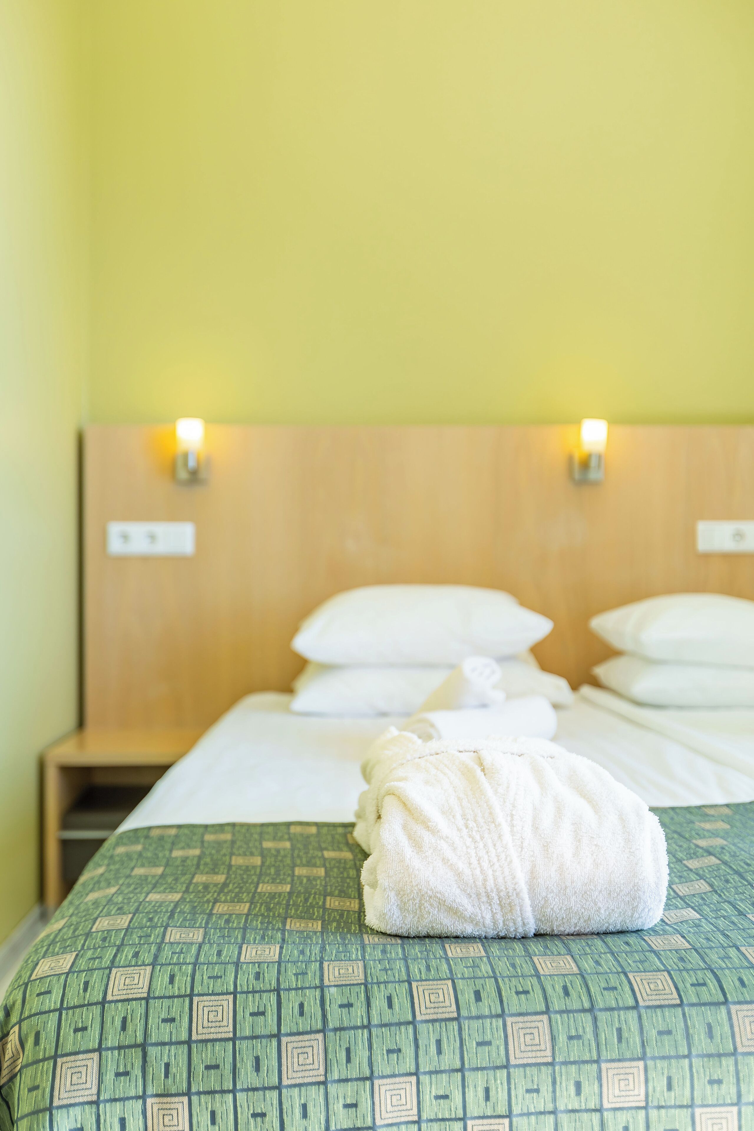Habitación de hotel con cabecera de madera, dos lámparas fijadas a la pared, cama con almohadas blancas y una toalla blanca doblada sobre la cama.