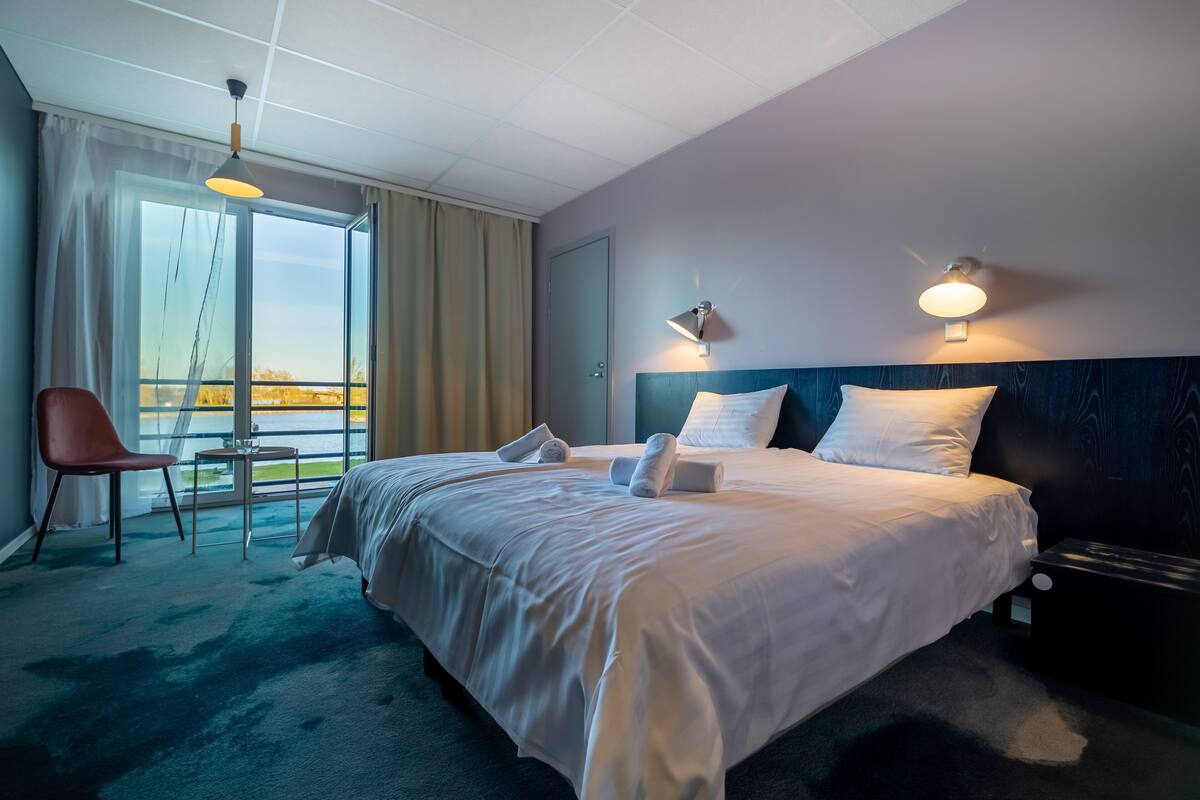 Ein Hotelzimmer mit einem großen Bett, einem Sessel, einem kleinen Tisch und Blick ins Freie.
